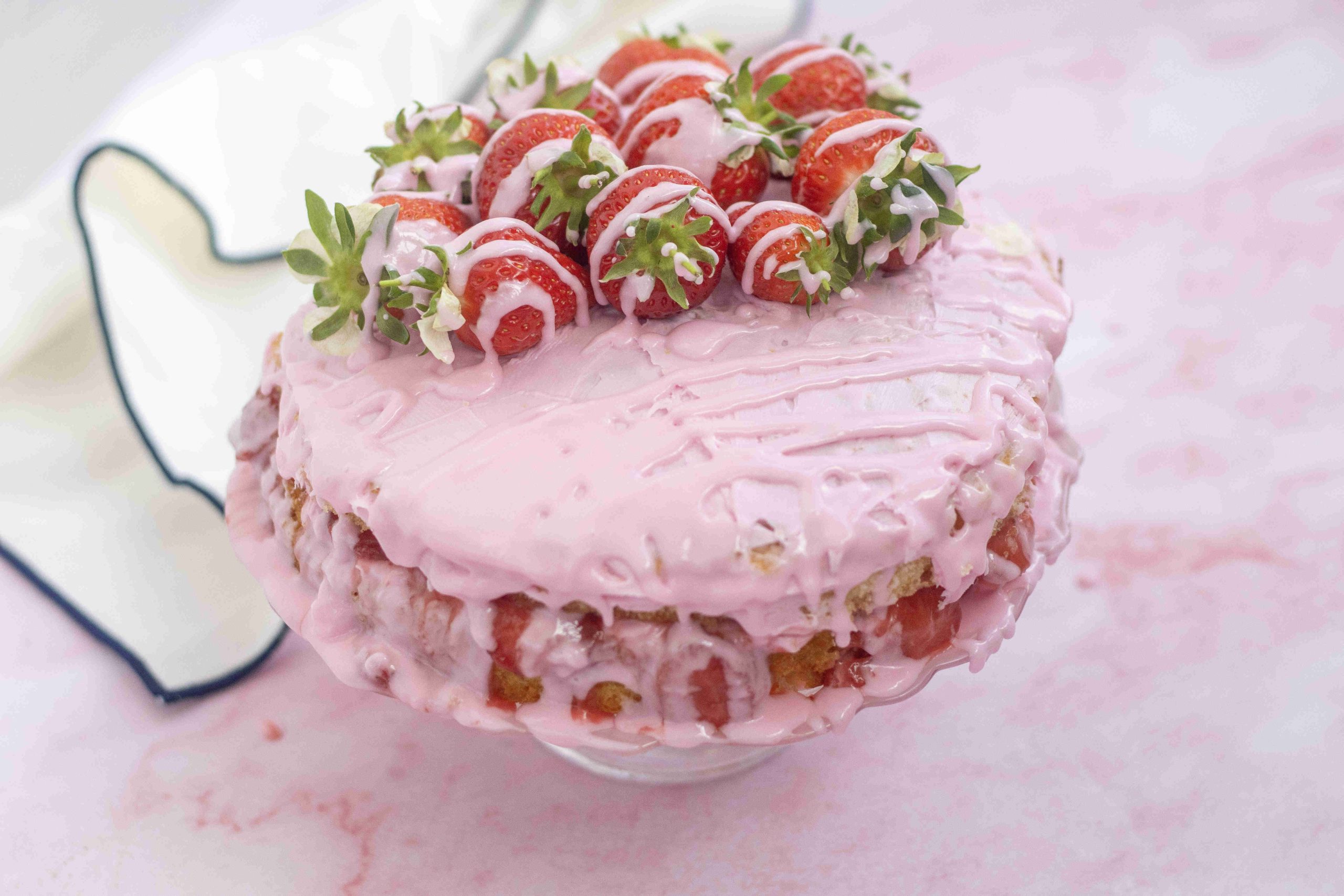 A pink frosted Valentine's Day cake topped with fresh strawberries and drizzled with white icing sits on a cake stand. Strawberry pieces show through its creamy, textured layers, with a white cloth in the background.