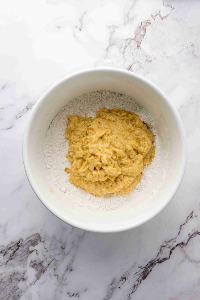 A white bowl on a marble surface containing flour and a thick yellow batter mixture being prepared for baking Christmas MM cookies.