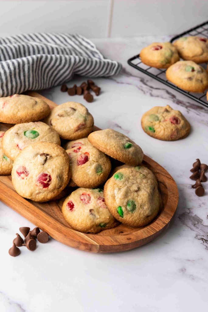 A wooden tray holds a pile of homemade Christmas MM cookies with red and green candy pieces. More cookies cool on a wire rack, with chocolate chips and a striped towel nearby on a marble countertop.