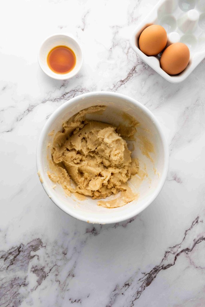 A white bowl with cookie dough for Christmas MM cookies sits on a marble surface. Nearby are a small bowl of vanilla extract and a carton holding two brown eggs.