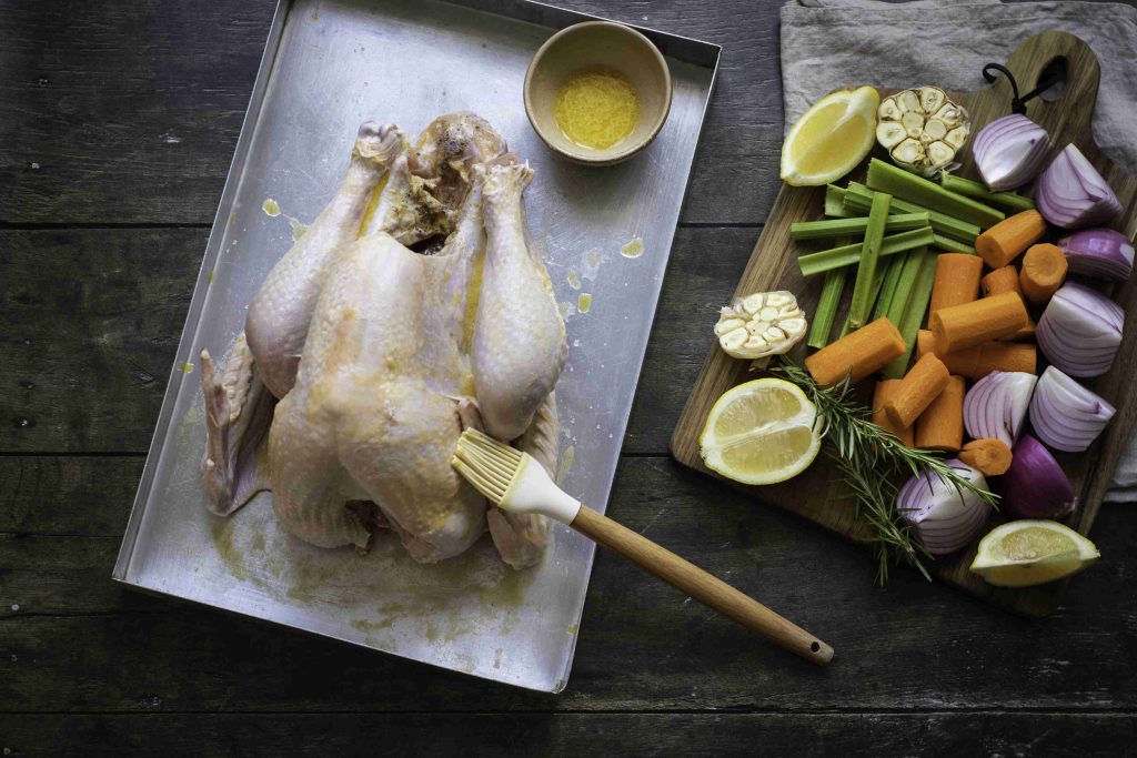 A raw whole chicken is brushed with seasoning on a metal tray beside a bowl of spices, evoking holiday turkey vibes. To the right, a wooden board holds cut lemons, garlic, celery, carrots, red onions, and rosemary on a rustic table.