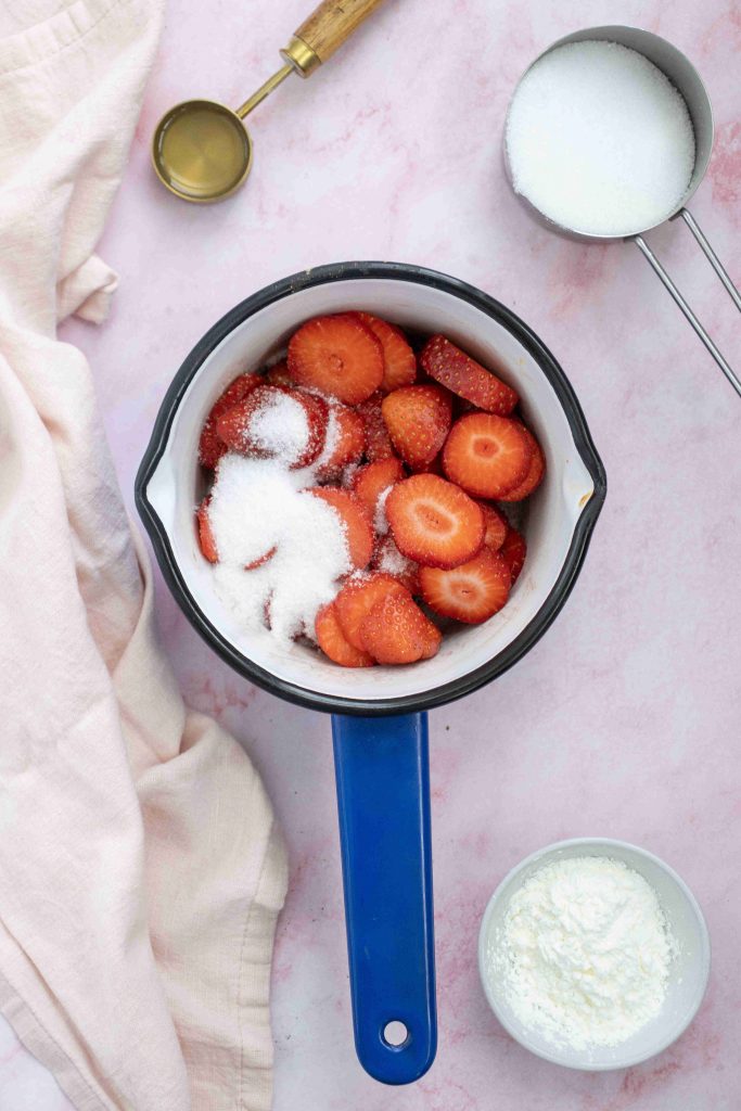 A blue saucepan filled with sliced strawberries and sugar, perfect for a Valentine's Day cake, is surrounded by a white measuring cup with sugar, a gold measuring spoon with liquid, a bowl of white powder, and a light pink cloth on a pink surface.