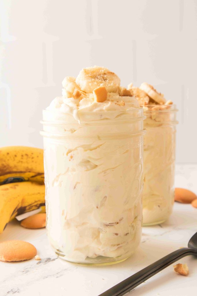 Two glass jars filled with creamy Christmas Banana Pudding, topped with banana slices and crushed vanilla wafers. In the background are ripe bananas and more vanilla wafer cookies on a white surface.
