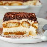 A slice of Christmas tiramisu on a white plate with a spoon, showing layers of creamy filling, coffee-soaked ladyfingers, and cocoa powder on top. A larger dish of Christmas tiramisu is in the background.