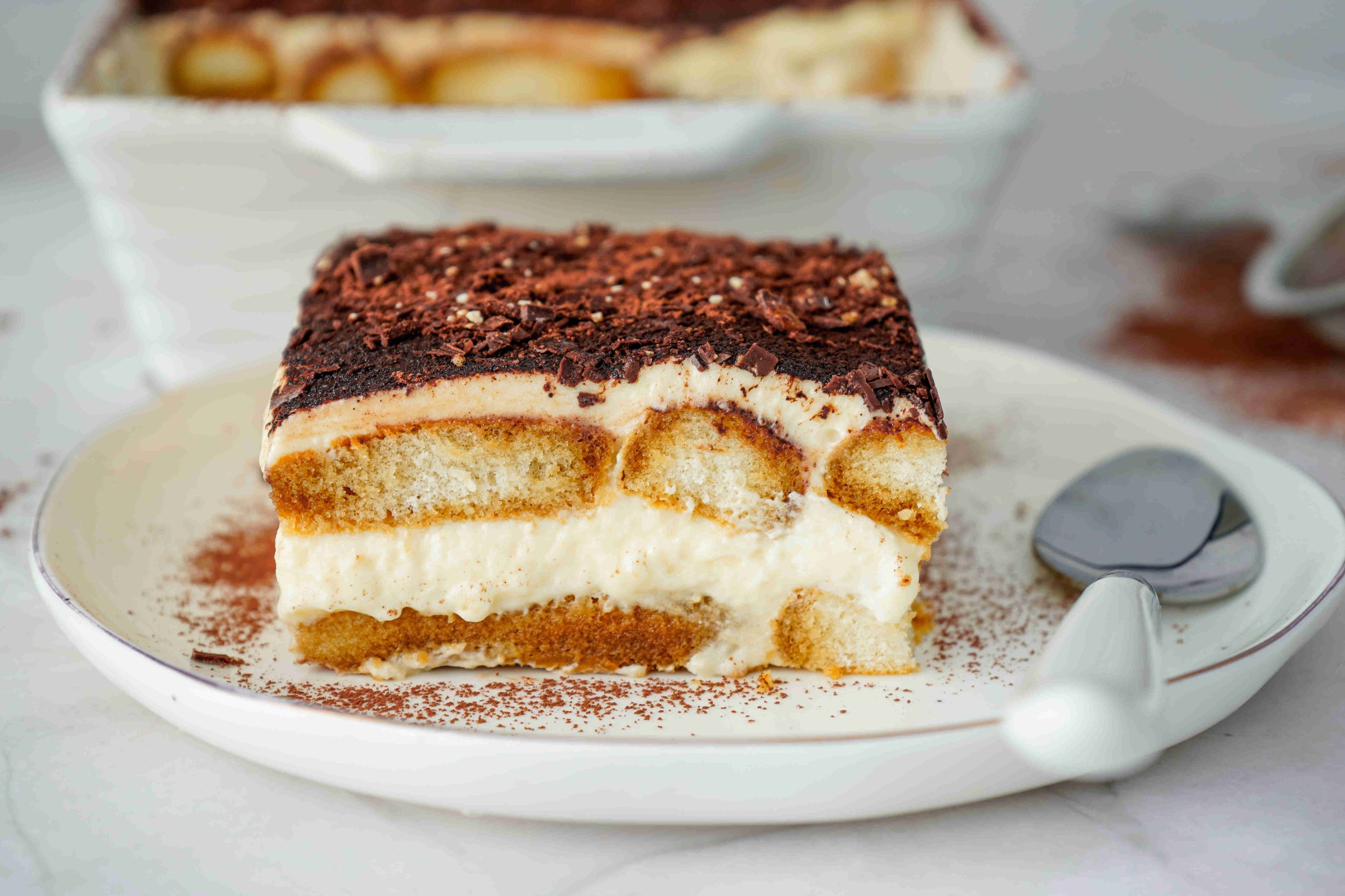 A slice of Christmas tiramisu on a white plate with a spoon, showing layers of creamy filling, coffee-soaked ladyfingers, and cocoa powder on top. A larger dish of Christmas tiramisu is in the background.