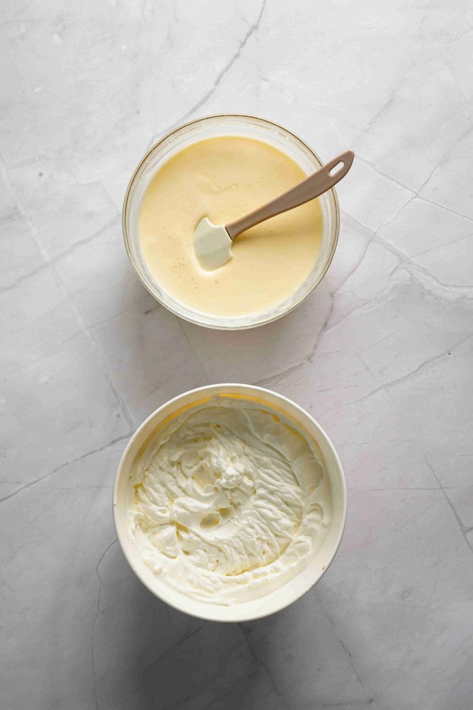 Two bowls on a marble surface: the top bowl holds a pale yellow, creamy mixture with a spatula, while the bottom bowl is filled with thick, white whipped cream—perfect layers for your Christmas tiramisu.