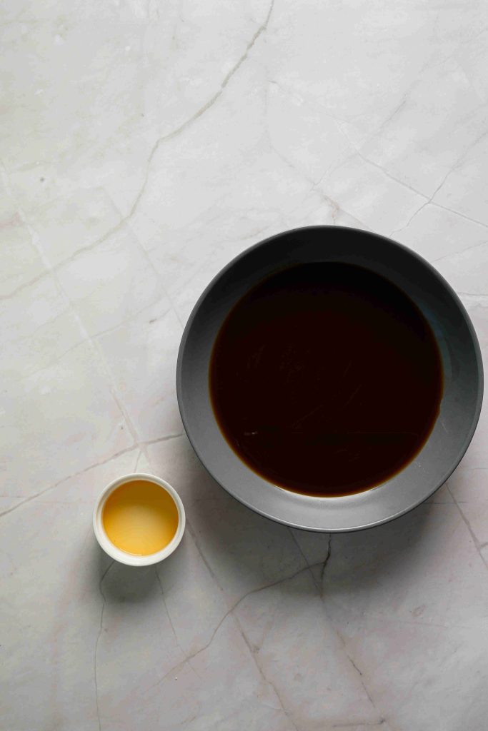 A dark grey bowl filled with a dark brown Christmas tiramisu mixture sits next to a small white cup containing a light yellow liquid, both placed on a white marble surface.