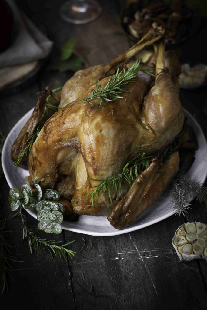A whole roasted Holiday Turkey garnished with fresh rosemary sits on a white plate, surrounded by herbs and a halved head of garlic on a rustic dark wooden table.