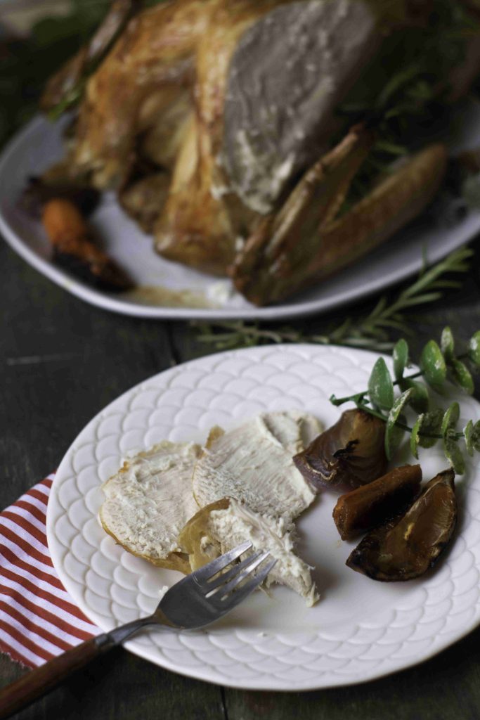 Sliced turkey breast and roasted vegetables on a white plate with a fork, in front of a whole Holiday Turkey on a separate plate, all set on a dark table with a red-striped napkin.