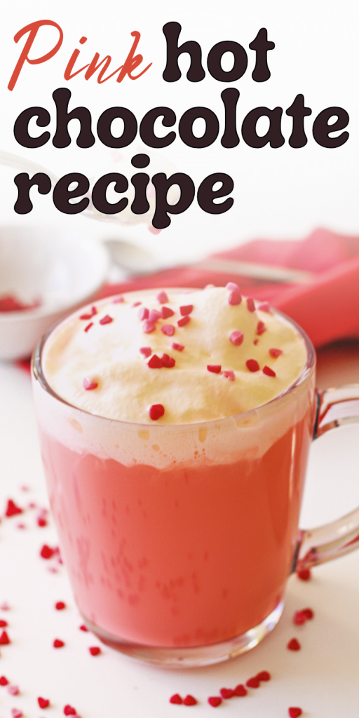 A glass mug filled with creamy pink hot chocolate, topped with whipped cream and red heart sprinkles—perfect for a Valentine’s Day hot chocolate treat. A red napkin and scattered sprinkles complete the scene, with “Pink hot chocolate recipe” in the background.