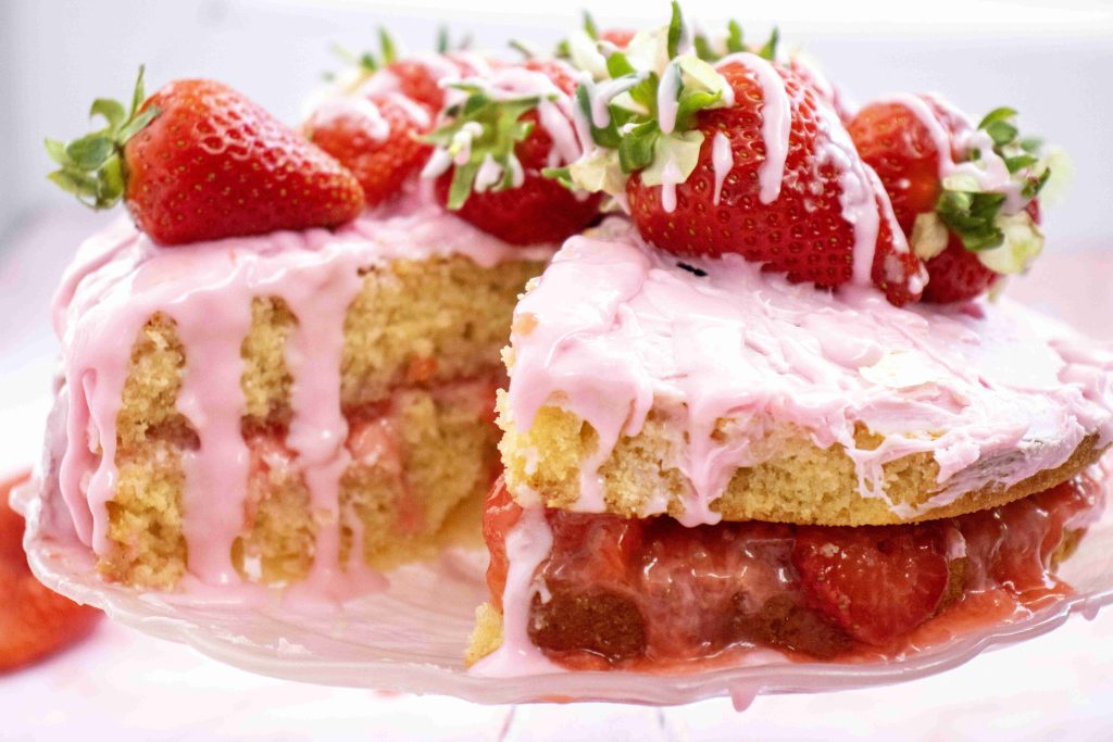 A layered strawberry valentine's day cake with pink icing and whole strawberries on top. A slice is slightly pulled out, showing the moist layers and strawberry filling inside. The cake sits on a clear glass plate.