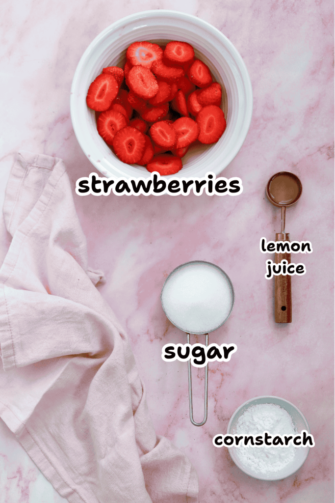 Flat lay of sliced strawberries in a white bowl for a valentine's day cake, with measuring cups of sugar, cornstarch, and lemon juice on a pink marble surface alongside a pale pink cloth. Each ingredient is labeled.