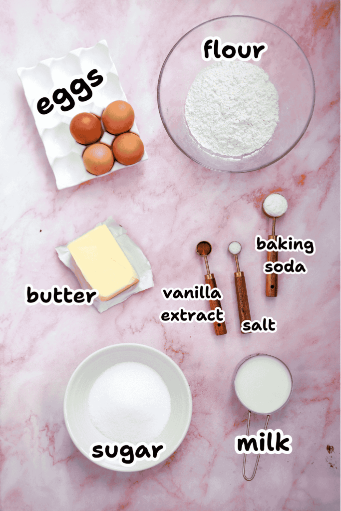 A flat lay of baking ingredients for a Valentine’s Day cake on a pink marble surface, labeled: eggs, flour, butter, vanilla extract, baking soda, salt, sugar, and milk.