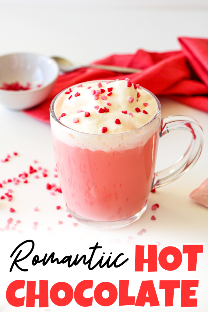 A glass mug of pink hot chocolate topped with whipped cream and red heart-shaped sprinkles sits on a white surface with scattered sprinkles, a red napkin, and a spoon in the background. Text reads Valentine’s Day hot chocolate.