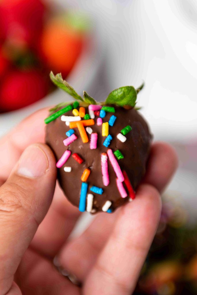 A hand holding a Valentine's Day chocolate covered strawberry topped with colorful rainbow sprinkles; blurred strawberries in the background.