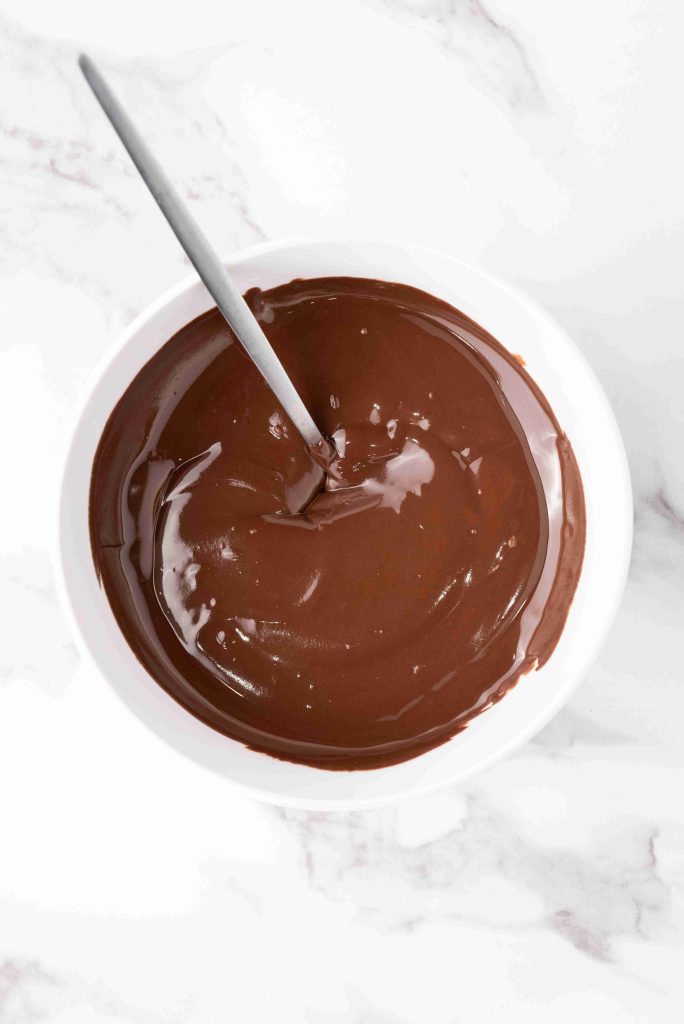 A white bowl filled with smooth, melted chocolate for valentine's day chocolate candy, with a metal spoon placed inside, on a white marble surface.