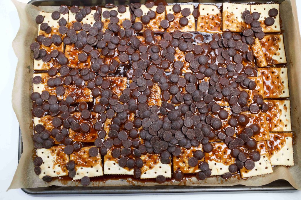 A baking tray lined with parchment paper holds saltine crackers topped with melted caramel and scattered chocolate chips—perfect for making Valentine’s Day Bark.
