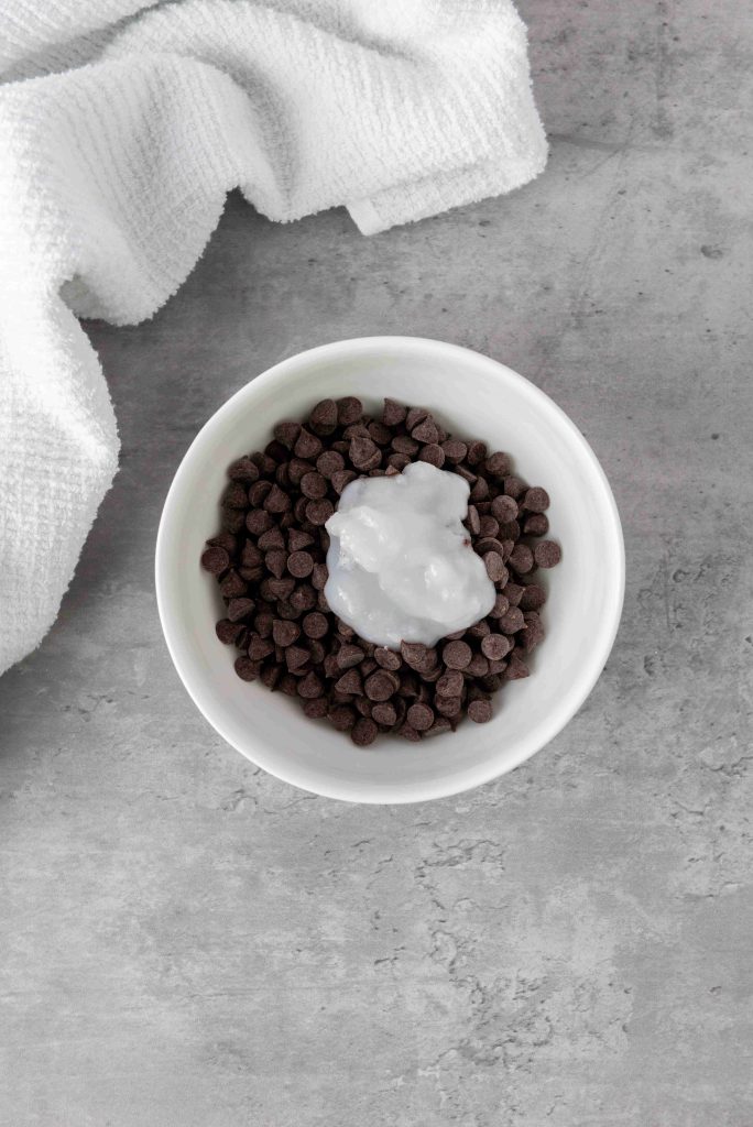 A white bowl filled with chocolate chips and a dollop of coconut oil, perfect for making Valentine's Day Chocolate Covered Strawberries, sits on a gray surface with a white textured cloth nearby.