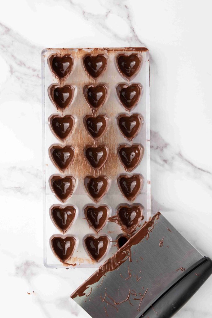 A heart-shaped chocolate mold filled with melted valentine's day chocolate candy sits on a white marble surface. A metal bench scraper with chocolate residue rests beside the mold.