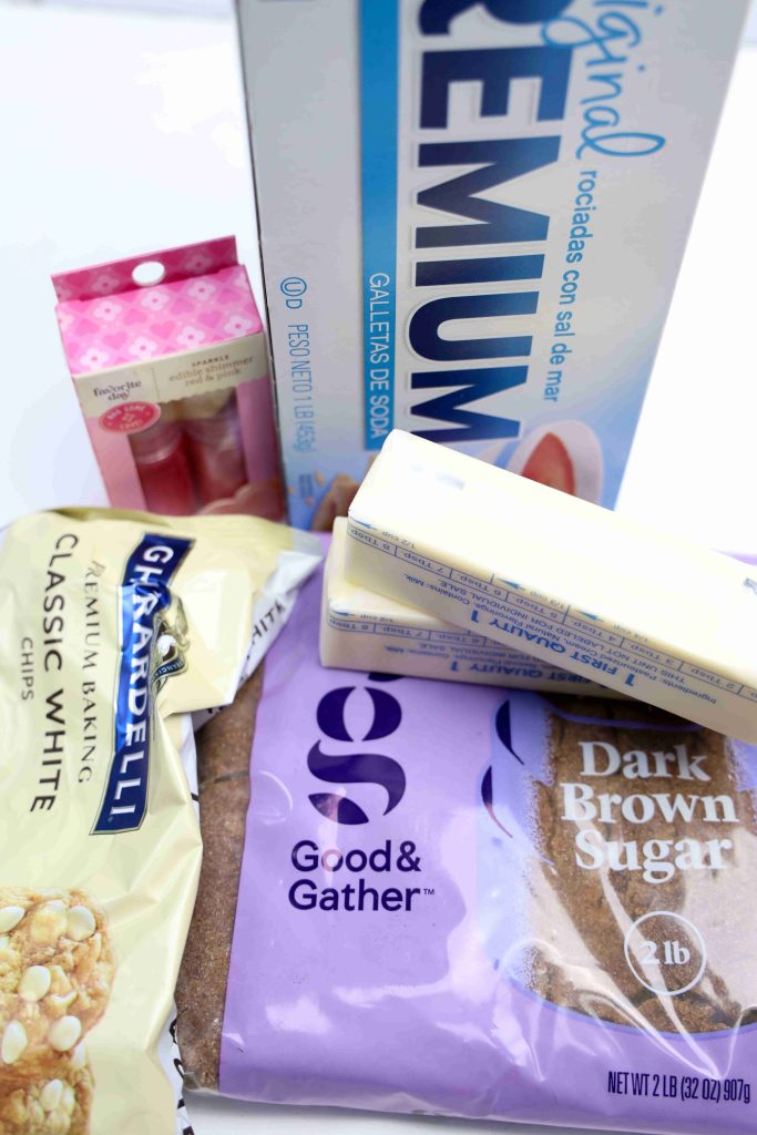 A close-up of baking ingredients for making Valentine’s Day Bark, featuring a box of saltine crackers, two sticks of butter, a package of dark brown sugar, white chocolate chips, and red food coloring.