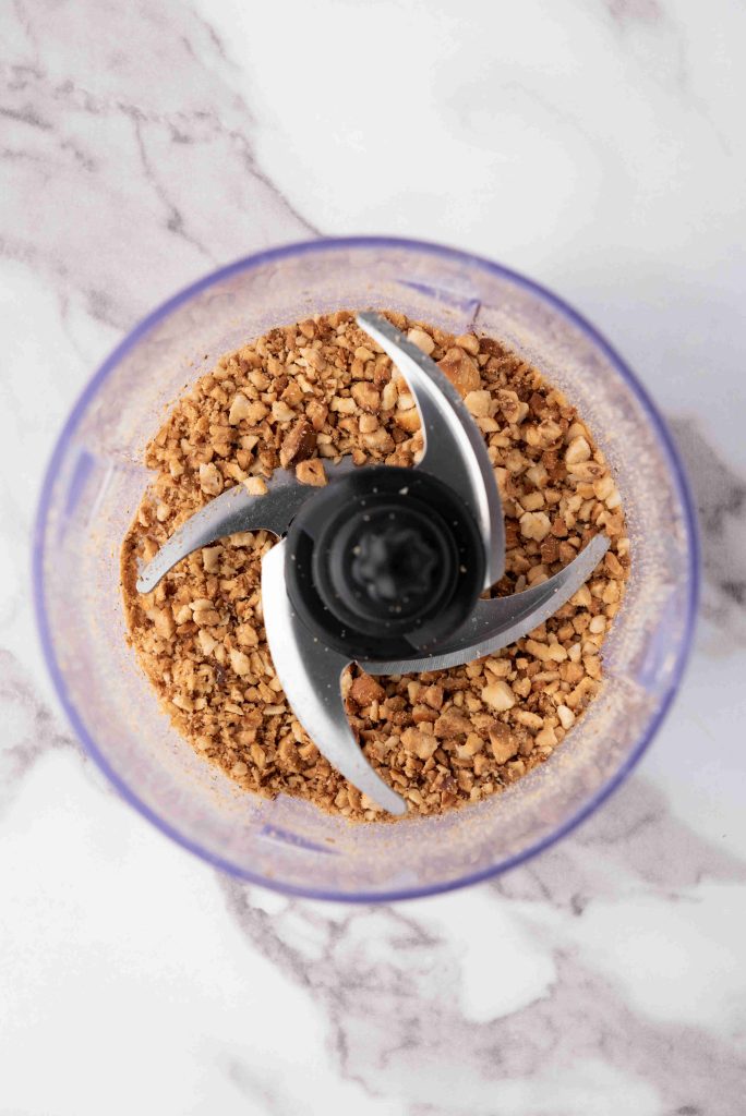 A food processor bowl with chopped nuts inside, perfect for making Valentine’s Day chocolate candy, viewed from above on a white marble surface.
