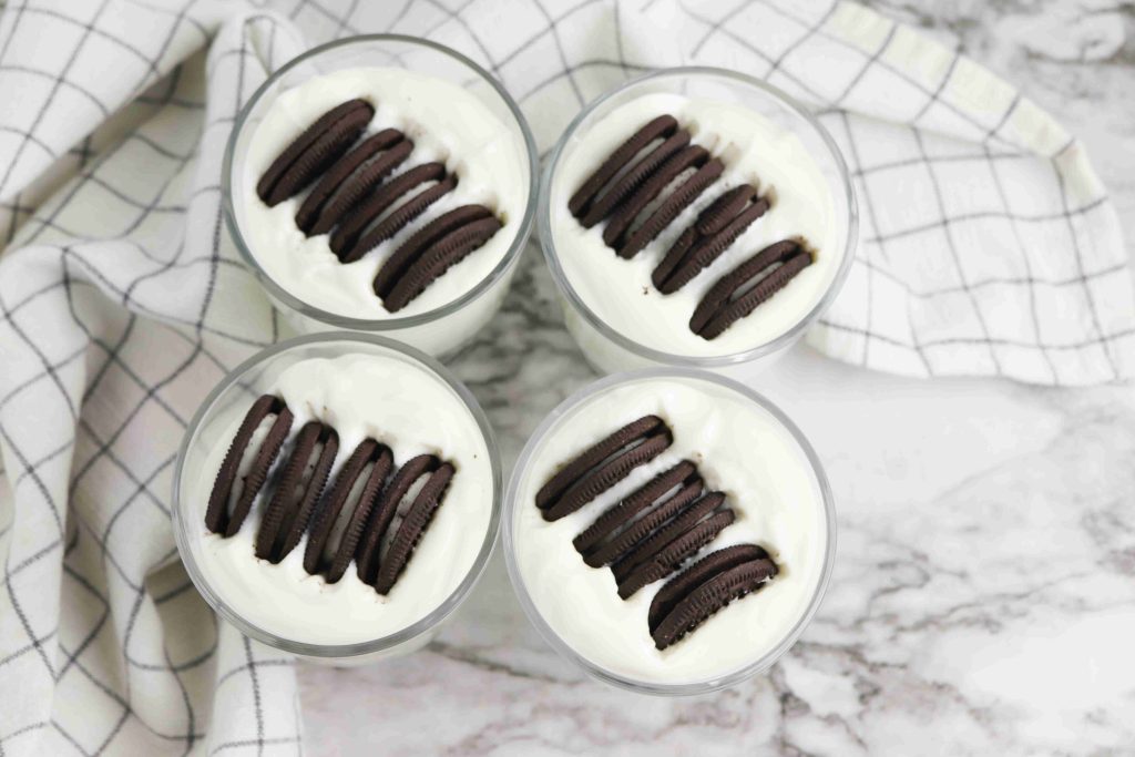 Four Oreo Yogurt Cups filled with creamy white dessert and topped with chocolate sandwich cookies, arranged on a marble surface with a white and gray checkered cloth.