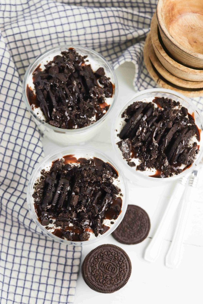 Three glass cups filled with creamy Oreo Yogurt Cups are topped with crushed chocolate sandwich cookies and chocolate drizzle. Two whole cookies and two white spoons sit beside the cups on a checkered cloth, near stacked wooden bowls.