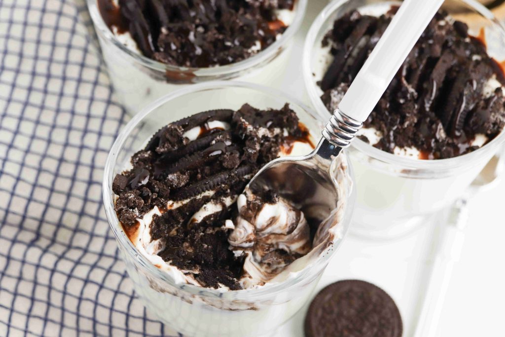 A glass filled with layers of whipped cream, crushed chocolate cookies, and chocolate syrup makes these Oreo Yogurt Cups irresistible. A spoon scoops some out; two more glasses and a whole cookie rest nearby on a checkered cloth.