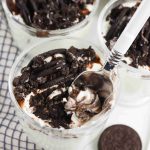 A glass filled with layers of whipped cream, crushed chocolate cookies, and chocolate syrup makes these Oreo Yogurt Cups irresistible. A spoon scoops some out; two more glasses and a whole cookie rest nearby on a checkered cloth.
