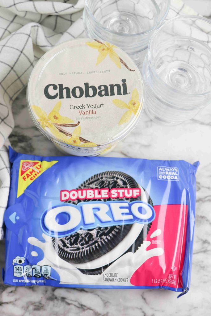 A container of Chobani vanilla Greek yogurt and a package of Double Stuf Oreo cookies rest on a marble surface, perfect for making delicious Oreo Yogurt Cups. Clear plastic bowls are stacked nearby for easy serving.