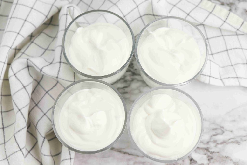 Four glass cups filled with plain white Oreo Yogurt Cups are arranged in a square on a marble surface, with a white and gray checkered cloth placed nearby.