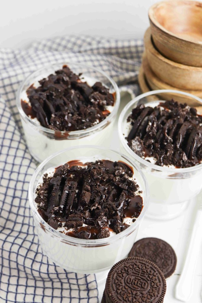 Three glass Oreo Yogurt Cups filled with creamy dessert, topped with crushed chocolate cookies and rich chocolate syrup, sit on a checkered cloth beside two whole sandwich cookies and wooden bowls.
