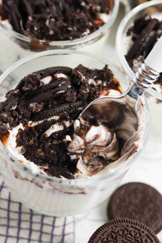 A close-up of Oreo Yogurt Cups filled with whipped cream, crushed chocolate cookies, and chocolate syrup, with a spoon scooping up a bite. A whole chocolate cookie and a checkered towel are nearby.