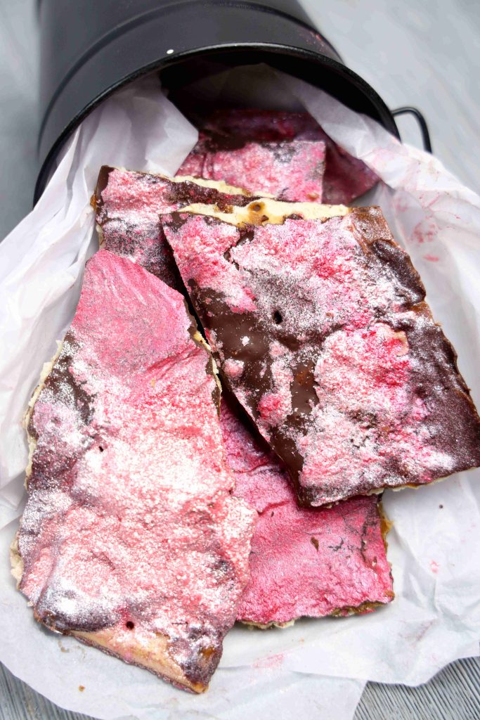 Several pieces of Valentine’s Day Bark matzo, covered in chocolate and a pink powdery topping, are arranged on white parchment paper inside a black container lying on its side.