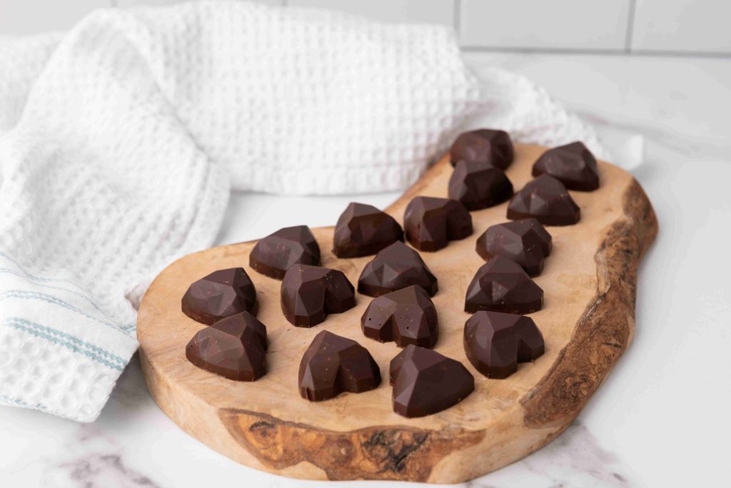 Heart-shaped dark chocolates, perfect as Valentine's Day chocolate candy, are arranged on a wooden serving board, with a white textured towel in the background on a marble countertop.