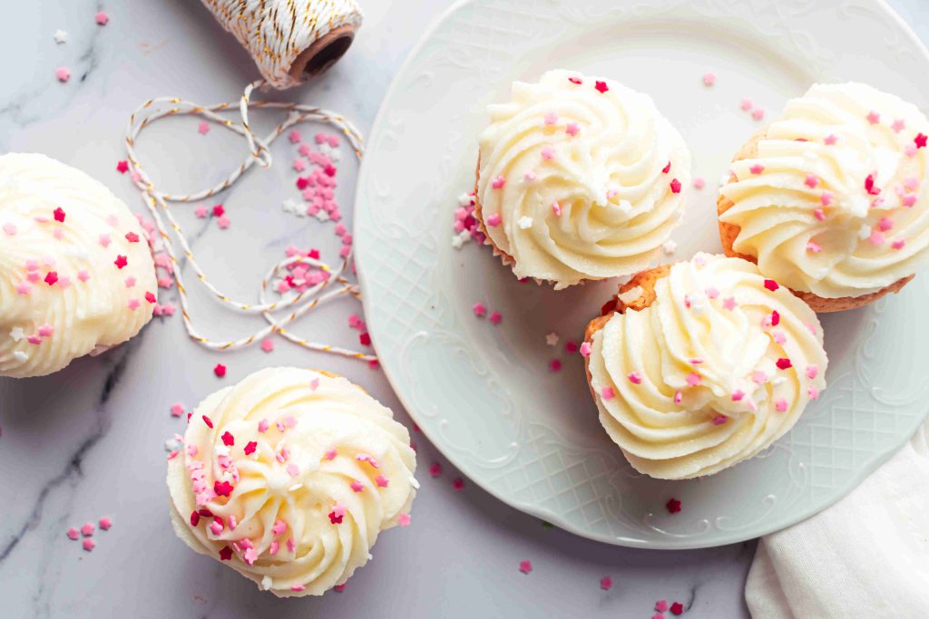 Four Valentine’s Day Cupcakes with swirled white frosting and pink sprinkles sit on a white plate and marble surface, with a spool of twine and scattered pink sprinkles nearby.
