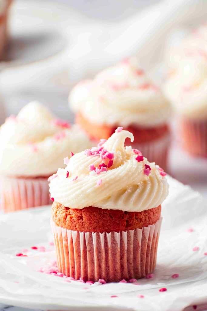 A close-up of a Valentine's Day Cupcake with white frosting and pink sprinkles on top, placed on a white surface. Other similarly decorated Valentine's Day cupcakes are blurred in the background.