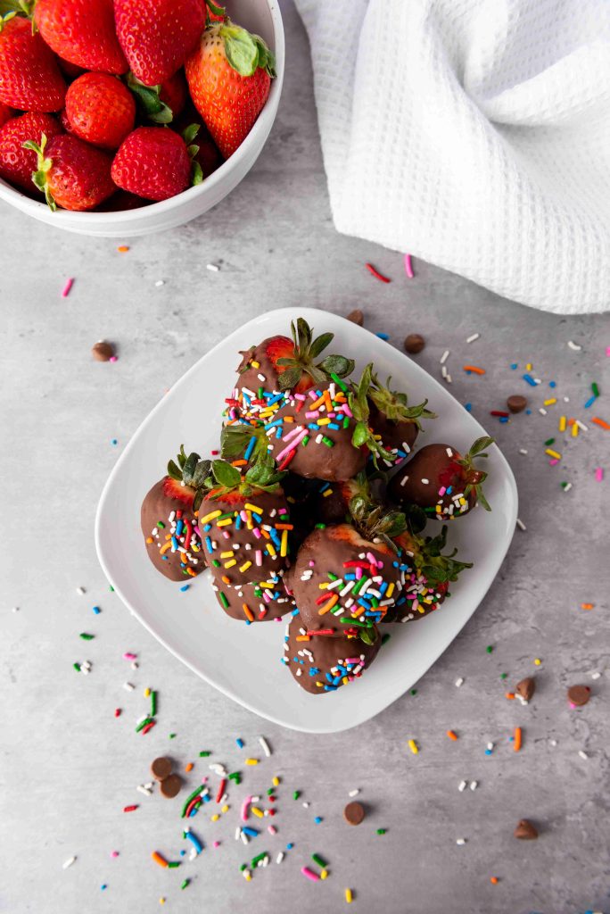 A plate of Valentine's Day Chocolate Covered Strawberries topped with colorful sprinkles sits on a gray surface. Nearby, a bowl of fresh strawberries and a white textured cloth add charm, with festive sprinkles scattered around.