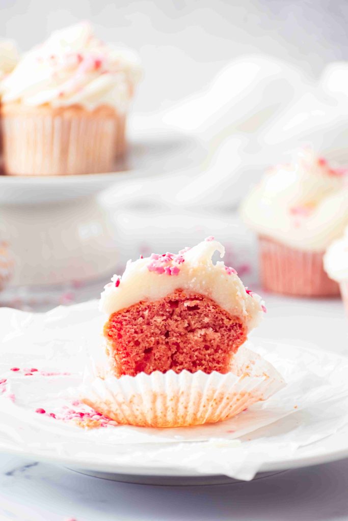 A partially eaten pink cupcake with white frosting and pink sprinkles sits on a white wrapper. More Valentine's Day Cupcakes, topped with festive frosting, are blurred in the background.