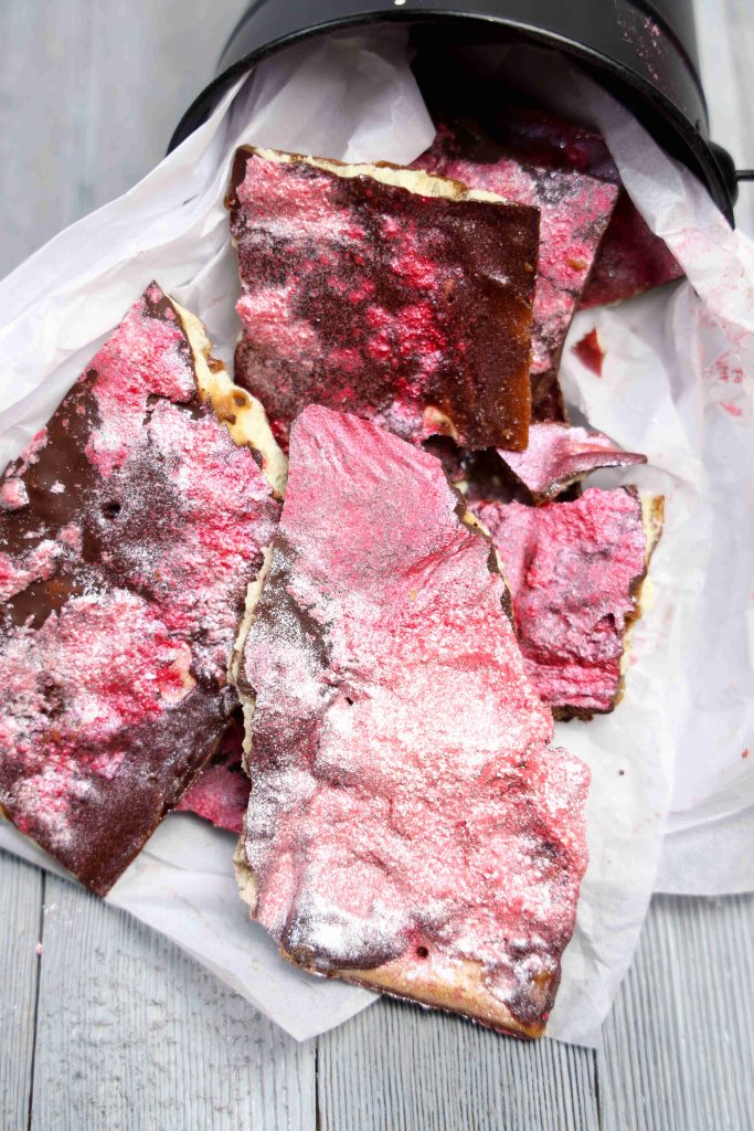 Valentine’s Day Bark pieces with a shiny, pink and purple metallic coating spill out of a black container lined with white parchment paper, set on a light wooden surface.
