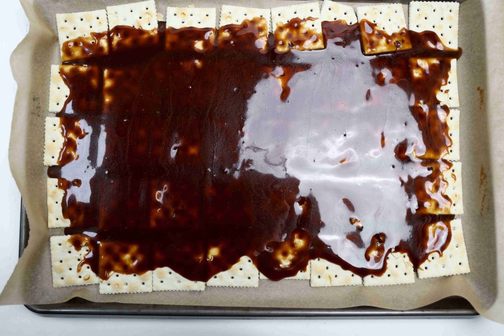 Saltine crackers lined up on a parchment-covered baking sheet, topped with a glossy layer of brown caramel sauce—this treat is the perfect base for Valentine’s Day Bark, halfway covering each cracker for a sweet and salty delight.