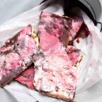 Several pieces of pink and chocolate-colored Valentine’s Day Bark are placed in a small metal bucket lined with parchment paper on a light wooden surface.