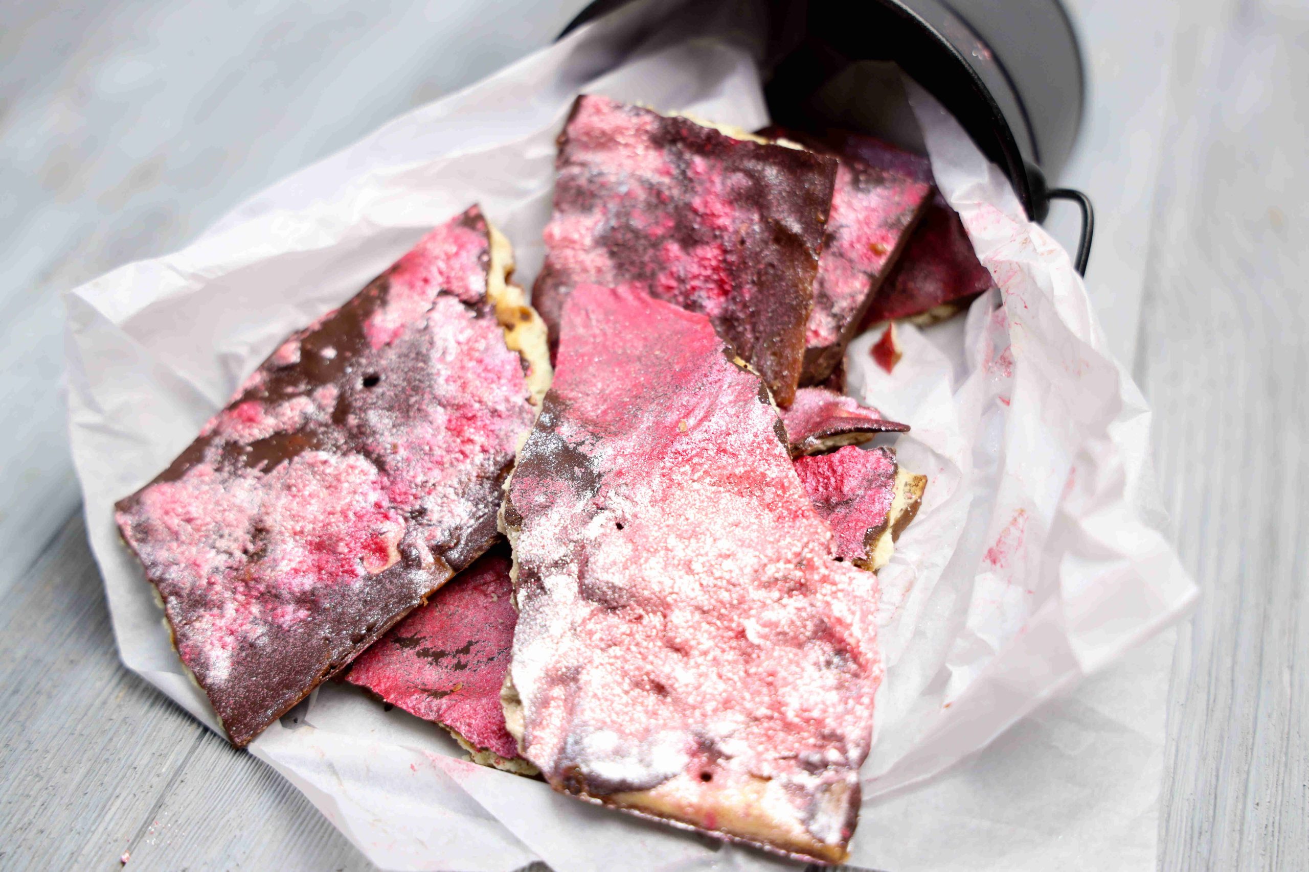 Several pieces of pink and chocolate-colored Valentine’s Day Bark are placed in a small metal bucket lined with parchment paper on a light wooden surface.