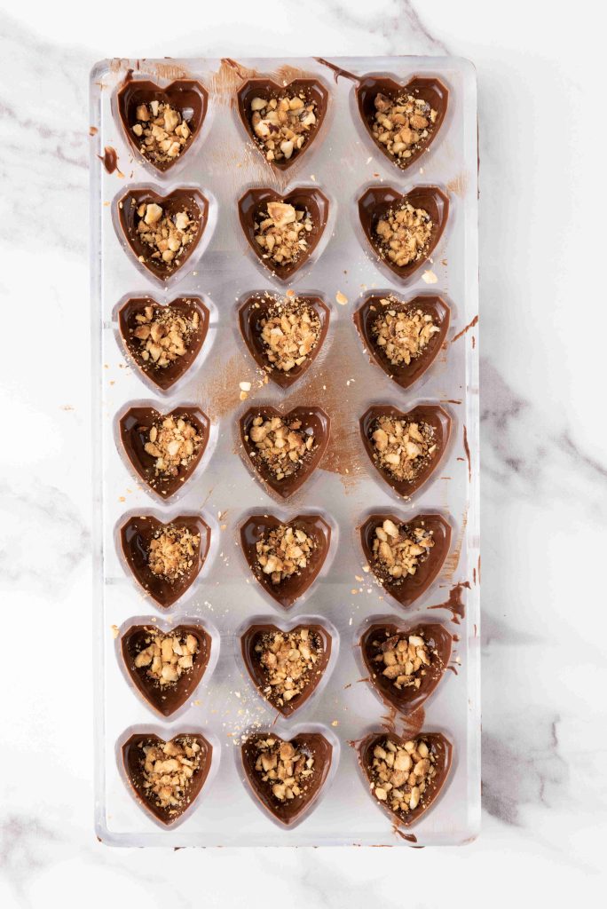 A clear plastic mold holds fifteen heart-shaped valentine's day chocolate candies, each filled with a crumbly nut mixture, placed on a white marble surface.