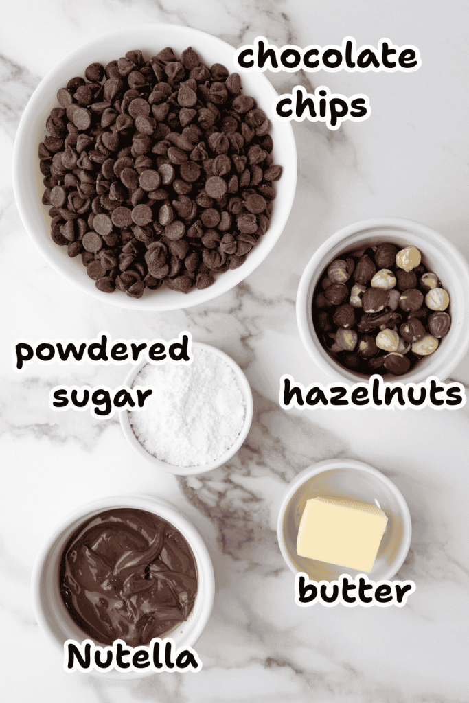 Five labeled bowls on a marble surface containing chocolate chips, hazelnuts, powdered sugar, butter, and Nutella—perfect ingredients for making Valentine’s Day chocolate candy—are beautifully arranged and viewed from above.