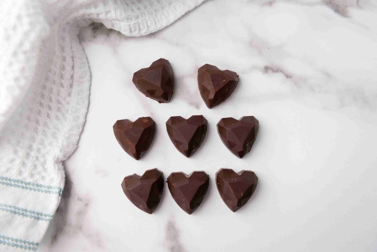 Eight heart-shaped dark chocolates, perfect as Valentine's Day chocolate candy, arranged in three rows on a white marble surface beside a white textured kitchen towel with blue stripes.