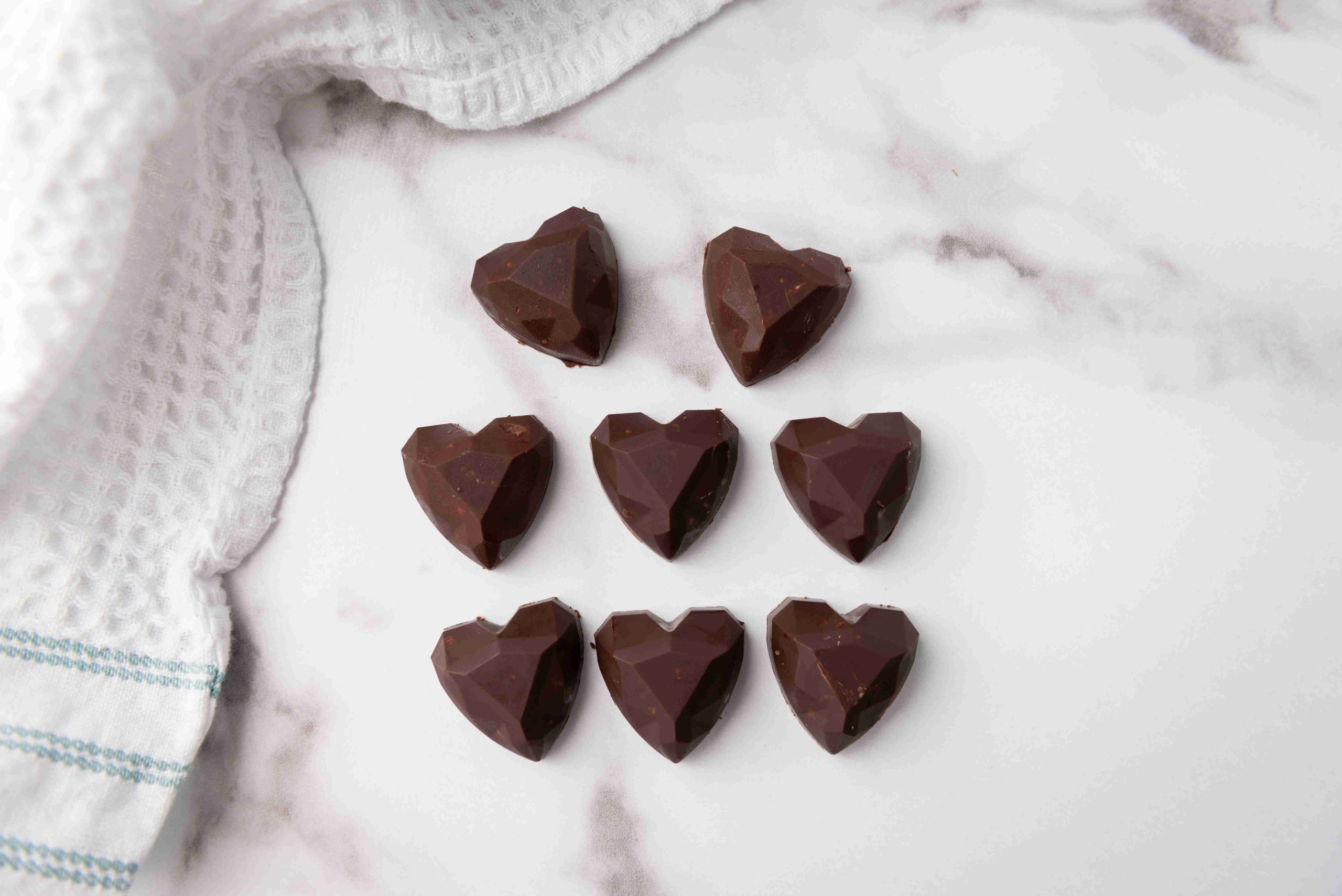Eight heart-shaped dark chocolates, perfect as Valentine's Day chocolate candy, arranged in three rows on a white marble surface beside a white textured kitchen towel with blue stripes.