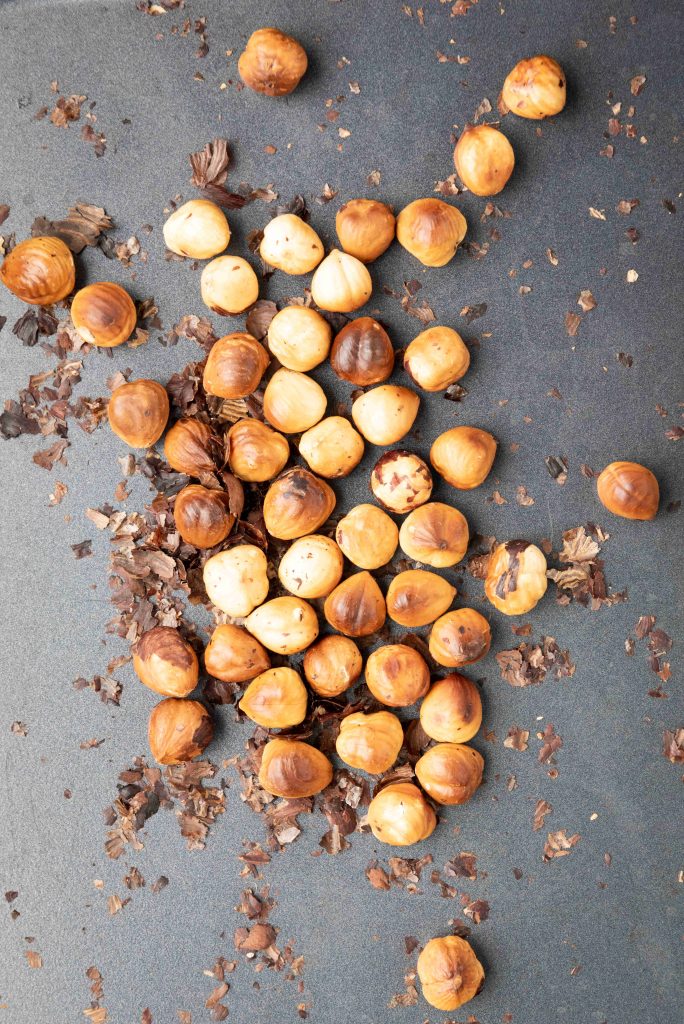 A scattered pile of roasted hazelnuts with brown skins and pieces of shell on a dark gray surface, perfect for making Valentine’s Day chocolate candy.