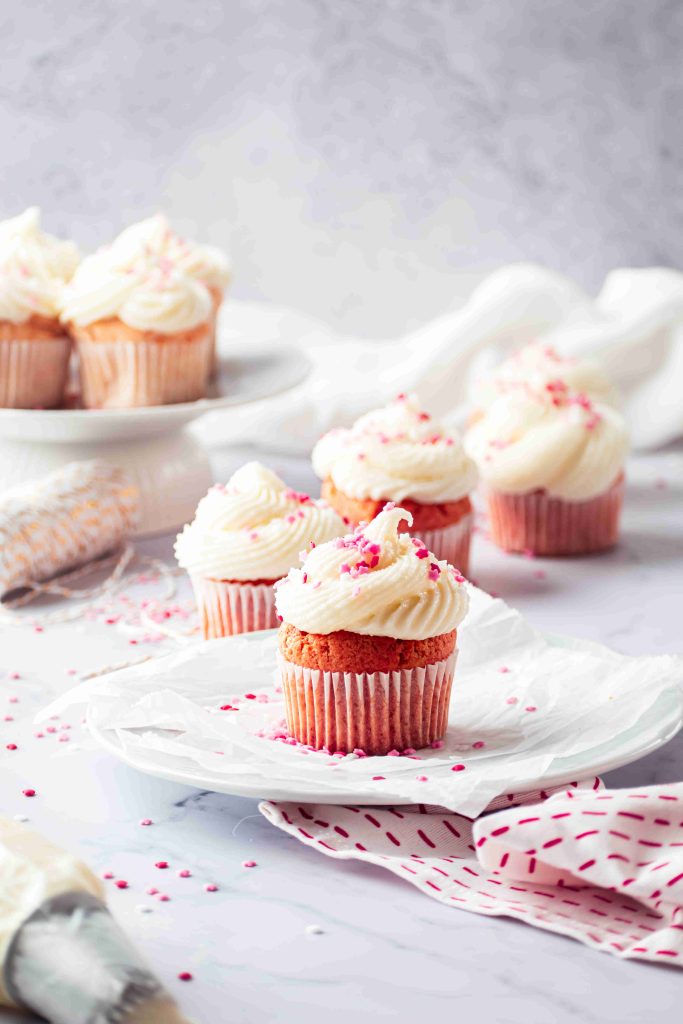 Valentine's Day Cupcakes with pink cake, white frosting, and pink sprinkles sit on a white plate atop a marble surface. More cupcakes and a piping bag are in the background, along with a white and pink cloth nearby.