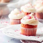 A close-up of a Valentine's Day cupcake with white frosting and pink sprinkles on a plate, with more decorated Valentine's Day cupcakes blurred in the background on a light, airy setting.